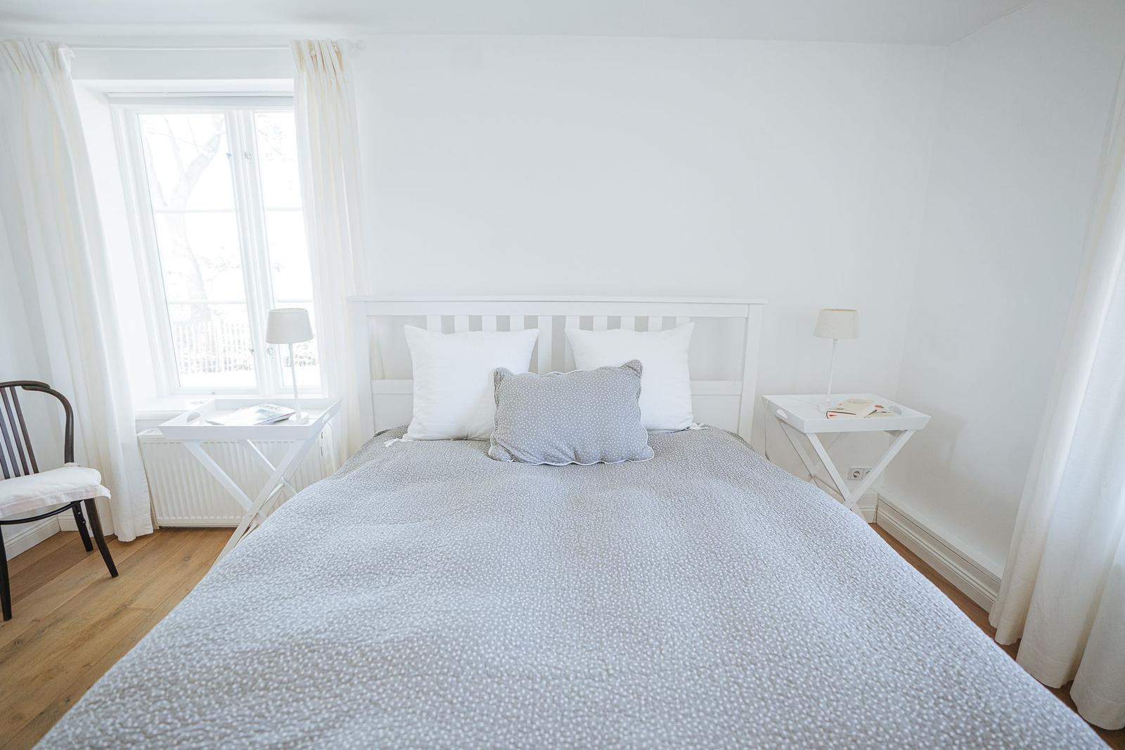 Schlafzimmer mit weißem Bett, zwei Nachttischen und Fenster mit weißen Vorhängen.