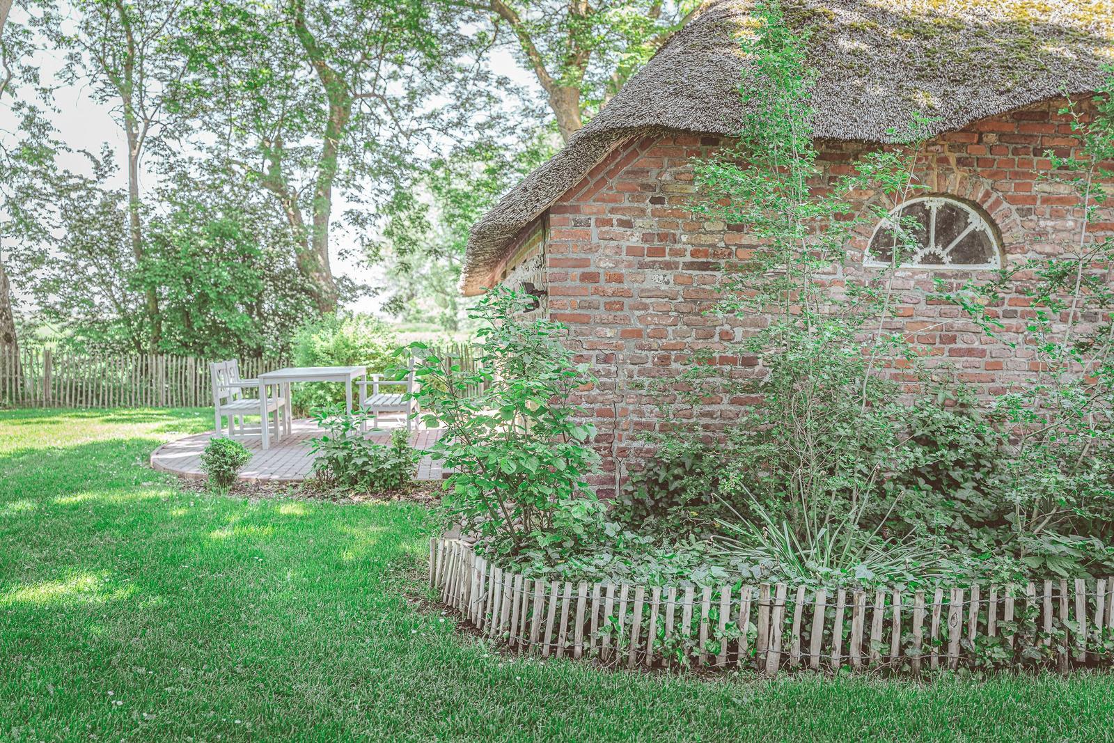 Haus mit Strohdach, weißer Tisch und Stühle auf Terrasse, umgeben von Gras und Bäumen.