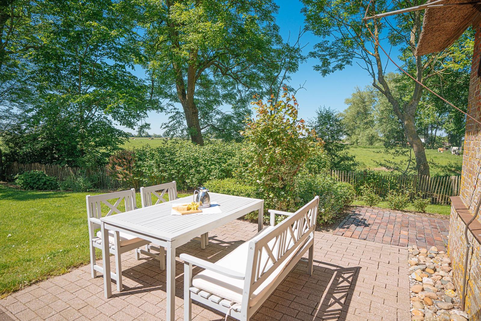 Terrasse mit weißem Tisch und Bänken, umgeben von Grün und Bäumen.
