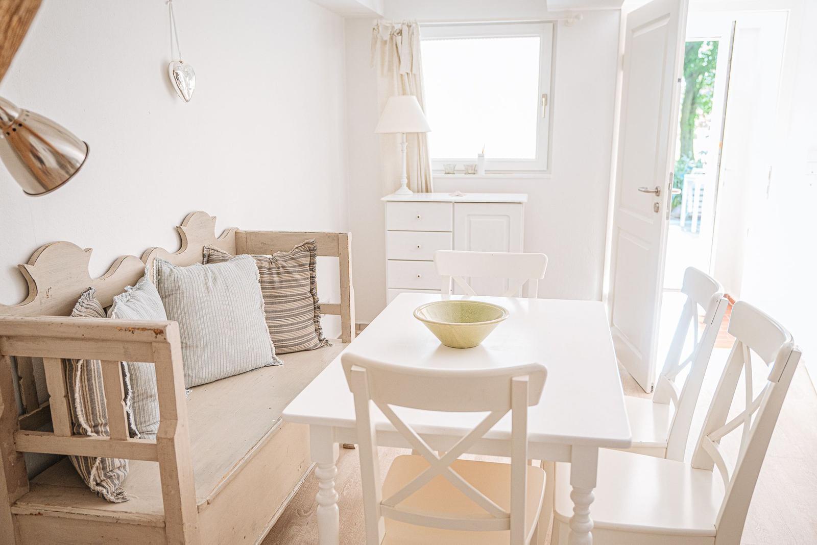 Esszimmer mit weißem Tisch, Stühlen und Sofa. Fenster und Tür führen nach draußen.