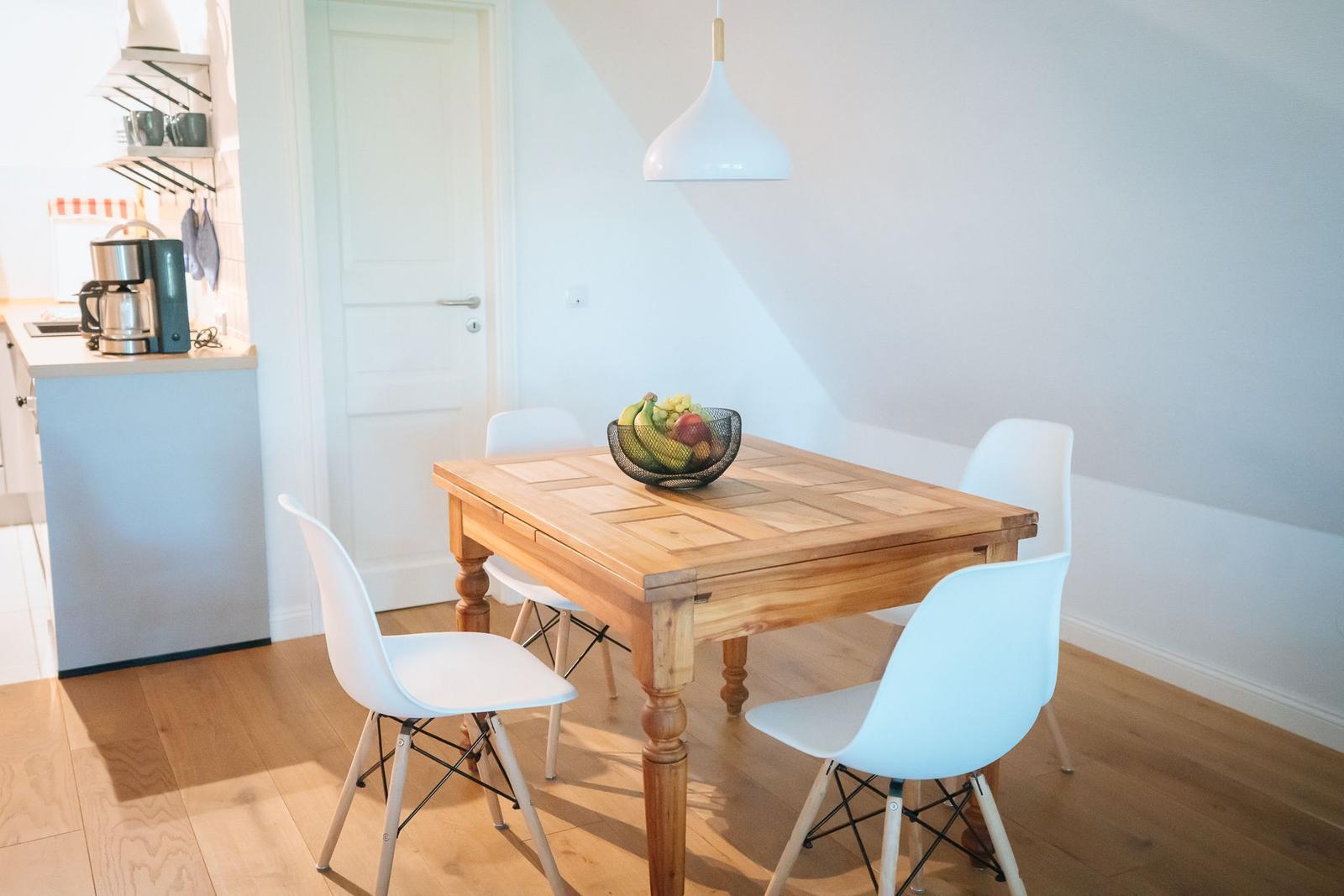 Essbereich mit Holztisch und weißen Stühlen. Küchenbereich im Hintergrund.