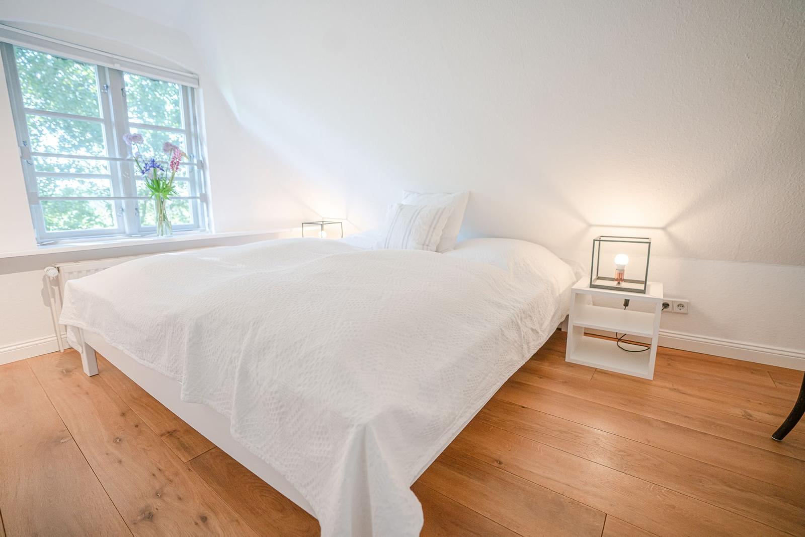 Schlafzimmer mit weißem Bett, Holzboden und Fenster mit Blumen.