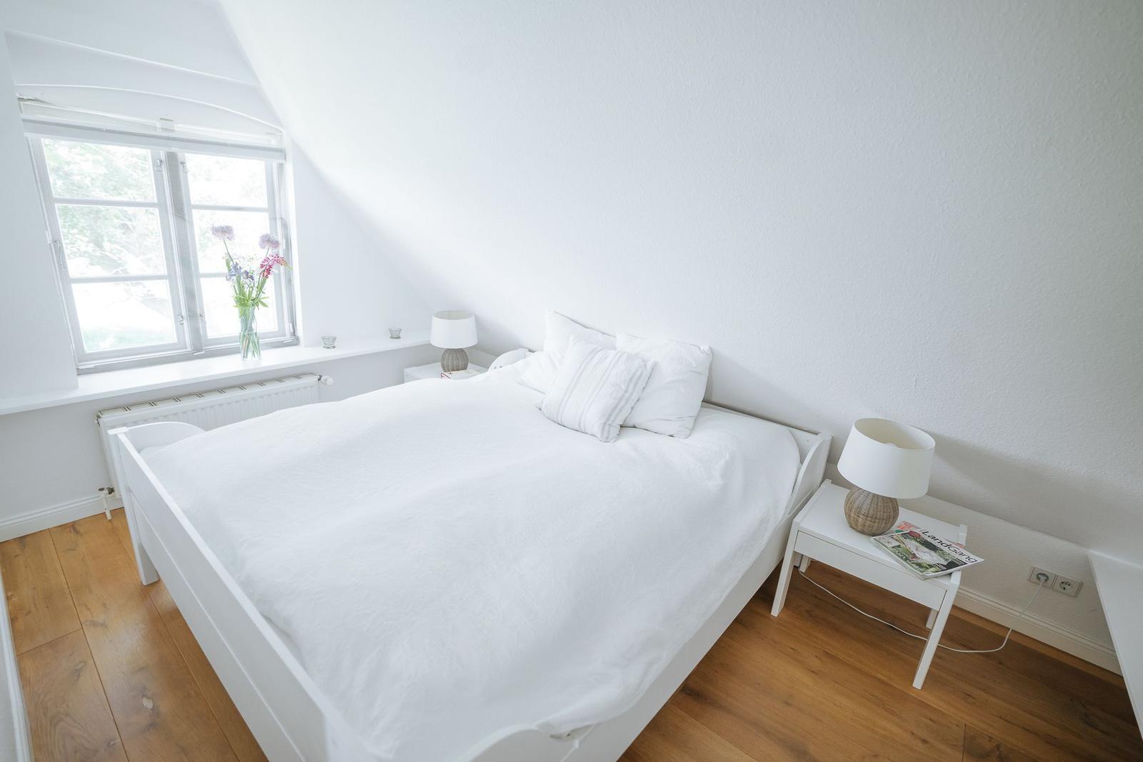 Schlafzimmer mit weißem Bett, Holzboden und Fenster mit Blumen.