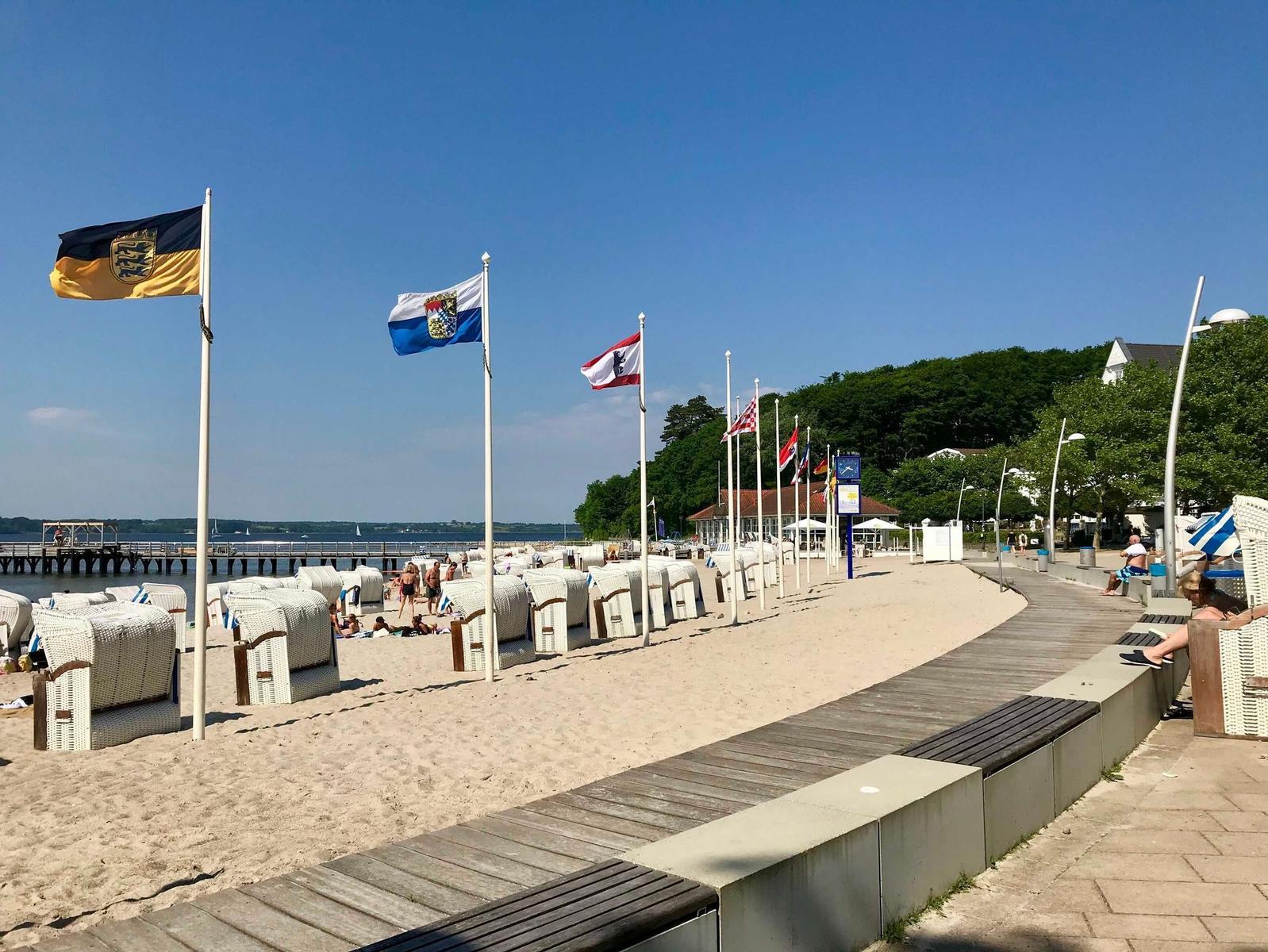 Strand mit Strandkörben, Holzweg und Flaggen am Ufer.