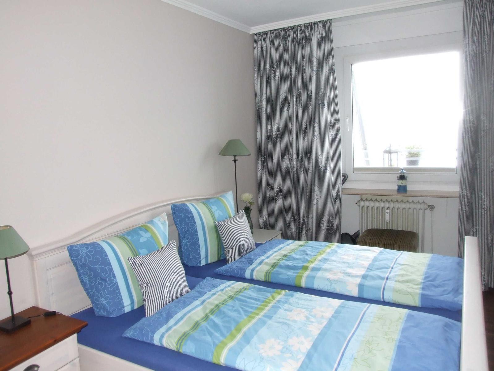 Zimmer mit Doppelbett, Fenster, Vorhängen und Nachttischen.