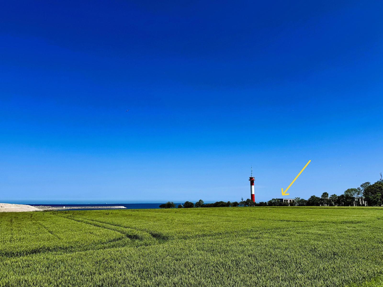 Rotes Leuchtturm mit weißem Streifen am Horizont über grüner Wiese und blauem Himmel.