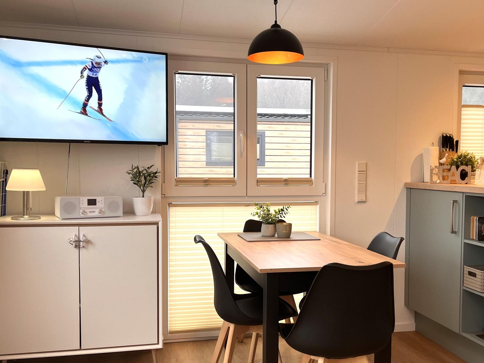 Essbereich mit Tisch, Stühlen und Fenster. TV zeigt Skifahrer. Küche daneben.