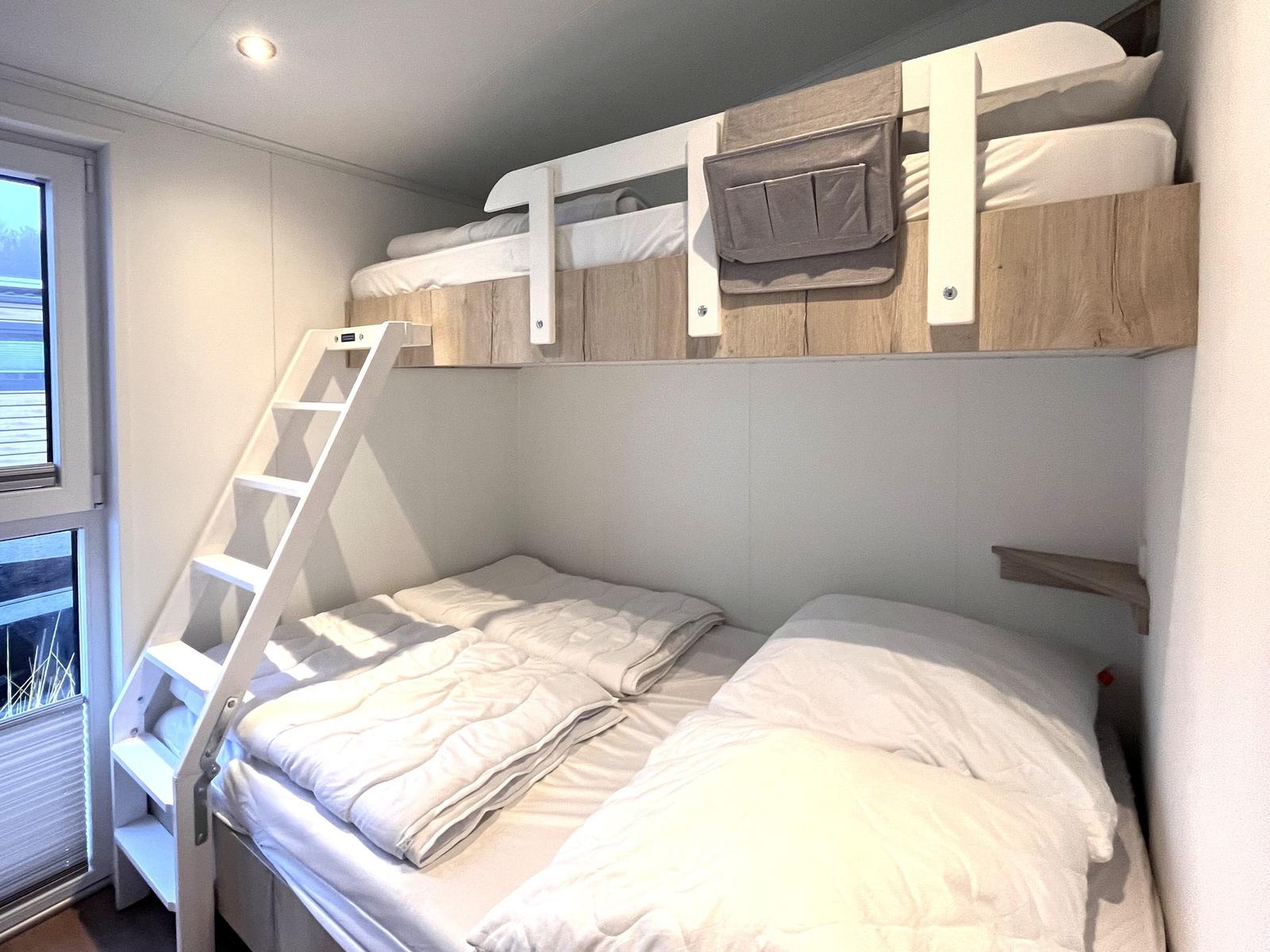 Zimmer mit Doppelbett und obere Schlafcouch, weiße Leiter und Fenster.