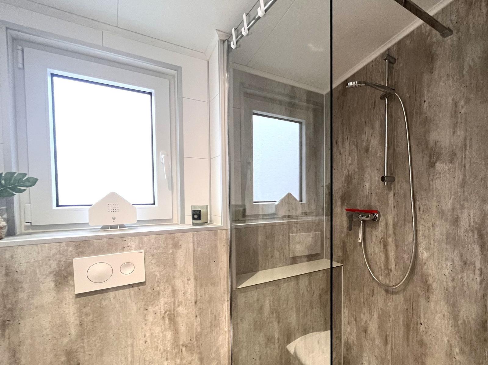 Badezimmer mit Dusche und Fenster. Betonwände und Glasduschtür.