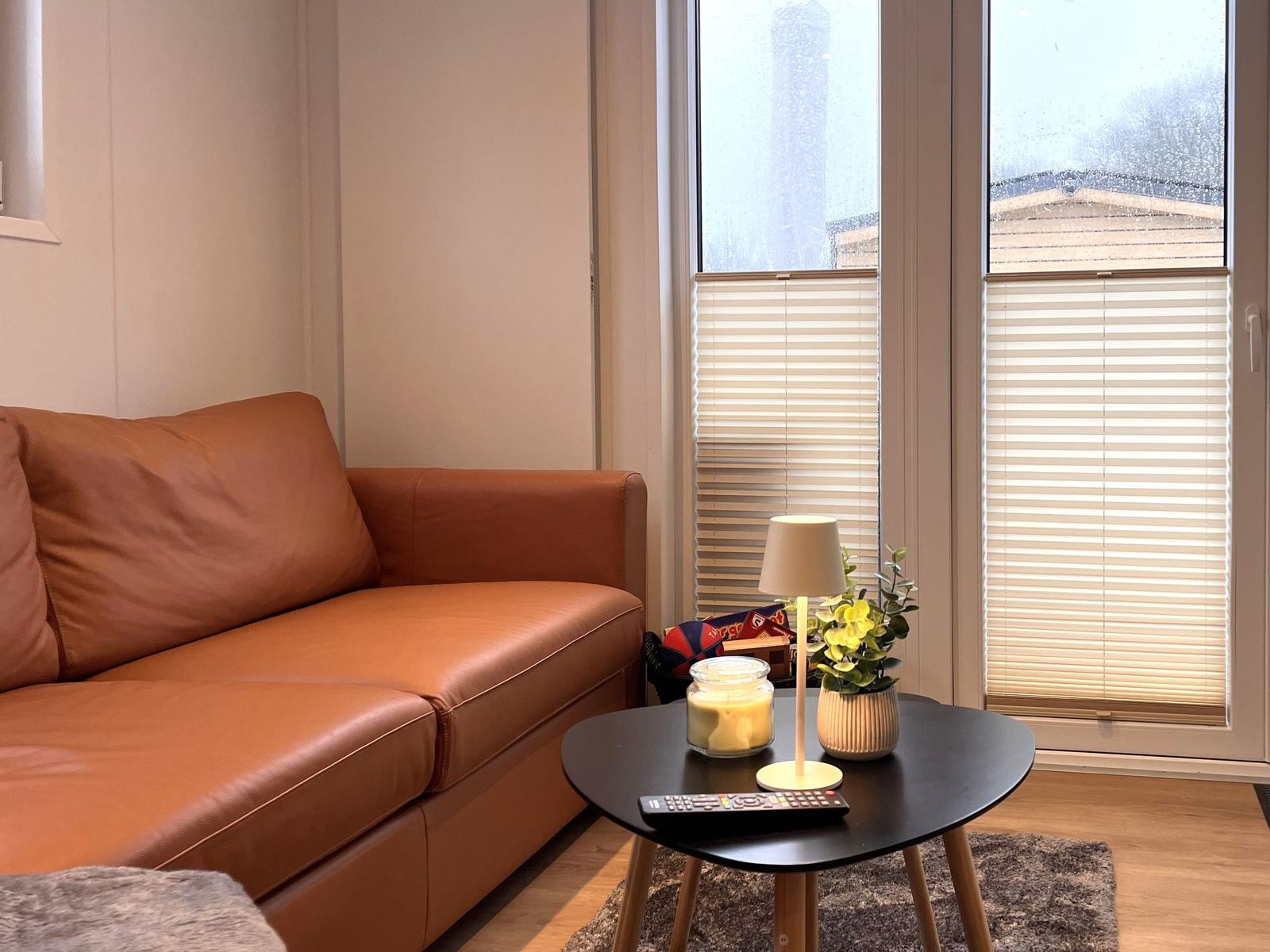 Ein braunes Ledersofa steht vor einem Fenster mit Jalousien. Davor steht ein runder Tisch mit Lampe und Pflanze.