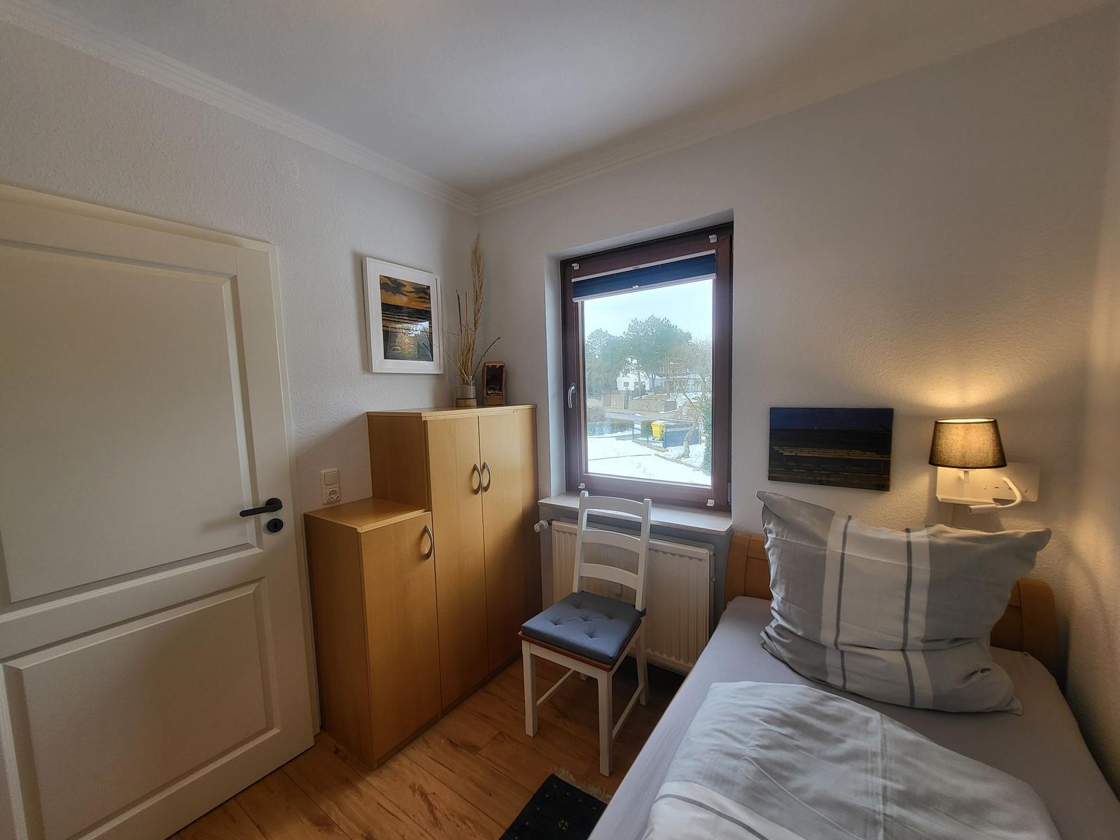 Schlafzimmer mit Bett, Schrank und Fenster. Wandlampe und Stuhl sind vorhanden.