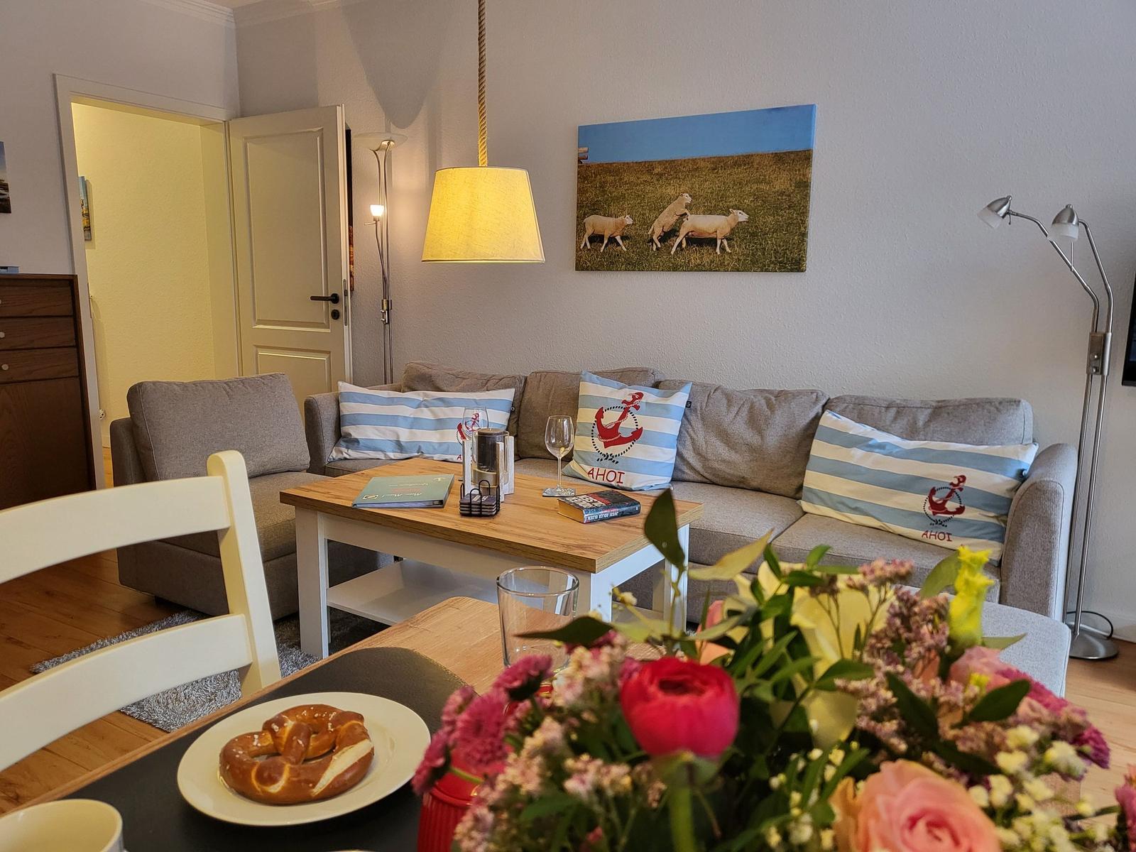 Wohnzimmer mit Sofa, Tisch und Blumen. Wandbild zeigt Schafe.