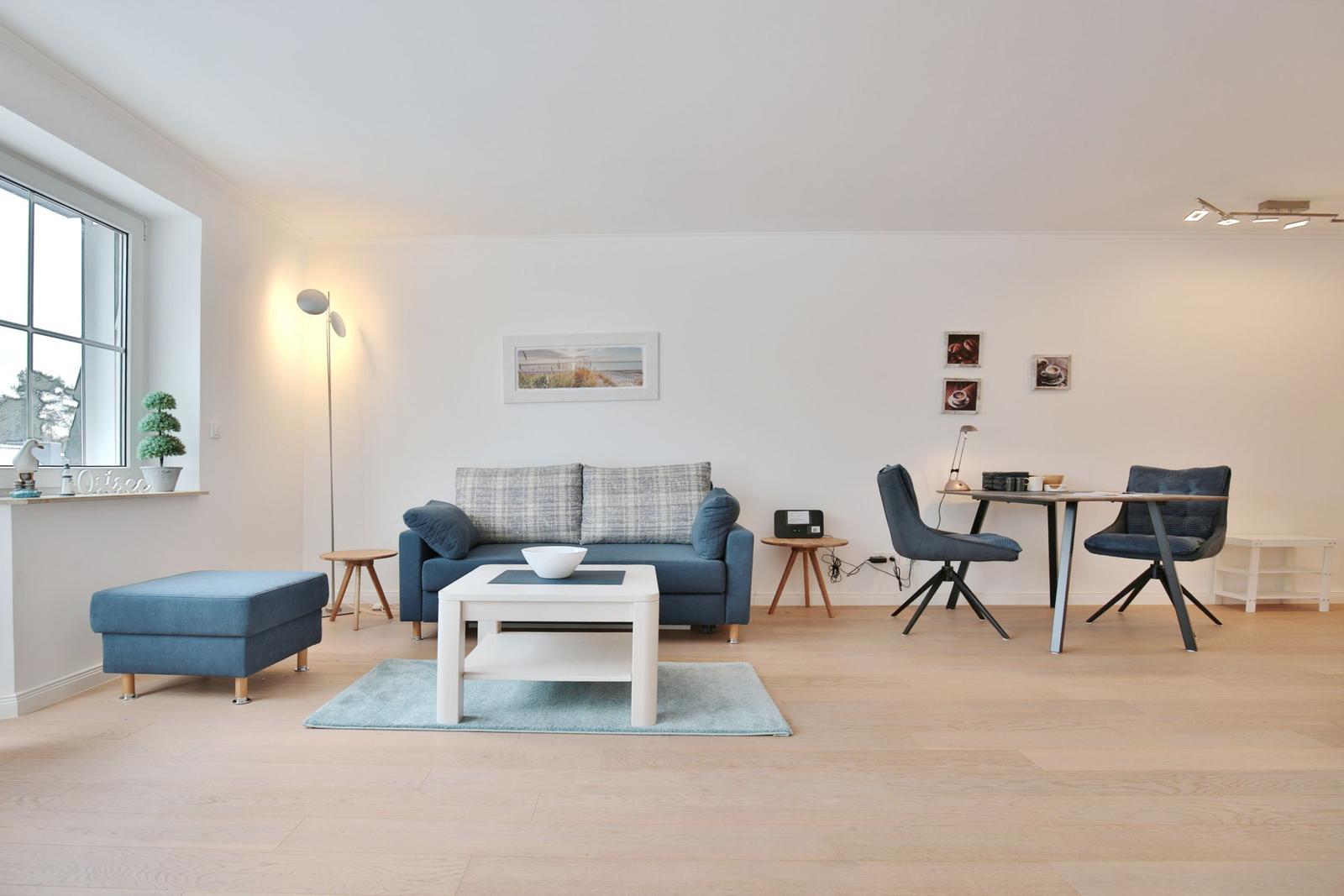 Wohnzimmer mit Sofa, Tisch und Stühlen. Holzboden und Fenster sichtbar.