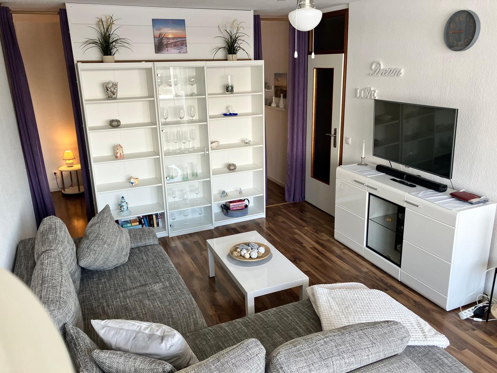 Wohnzimmer mit Sofa, Couchtisch und TV-Möbel. Hintergrund: Bücherregal und Tür.