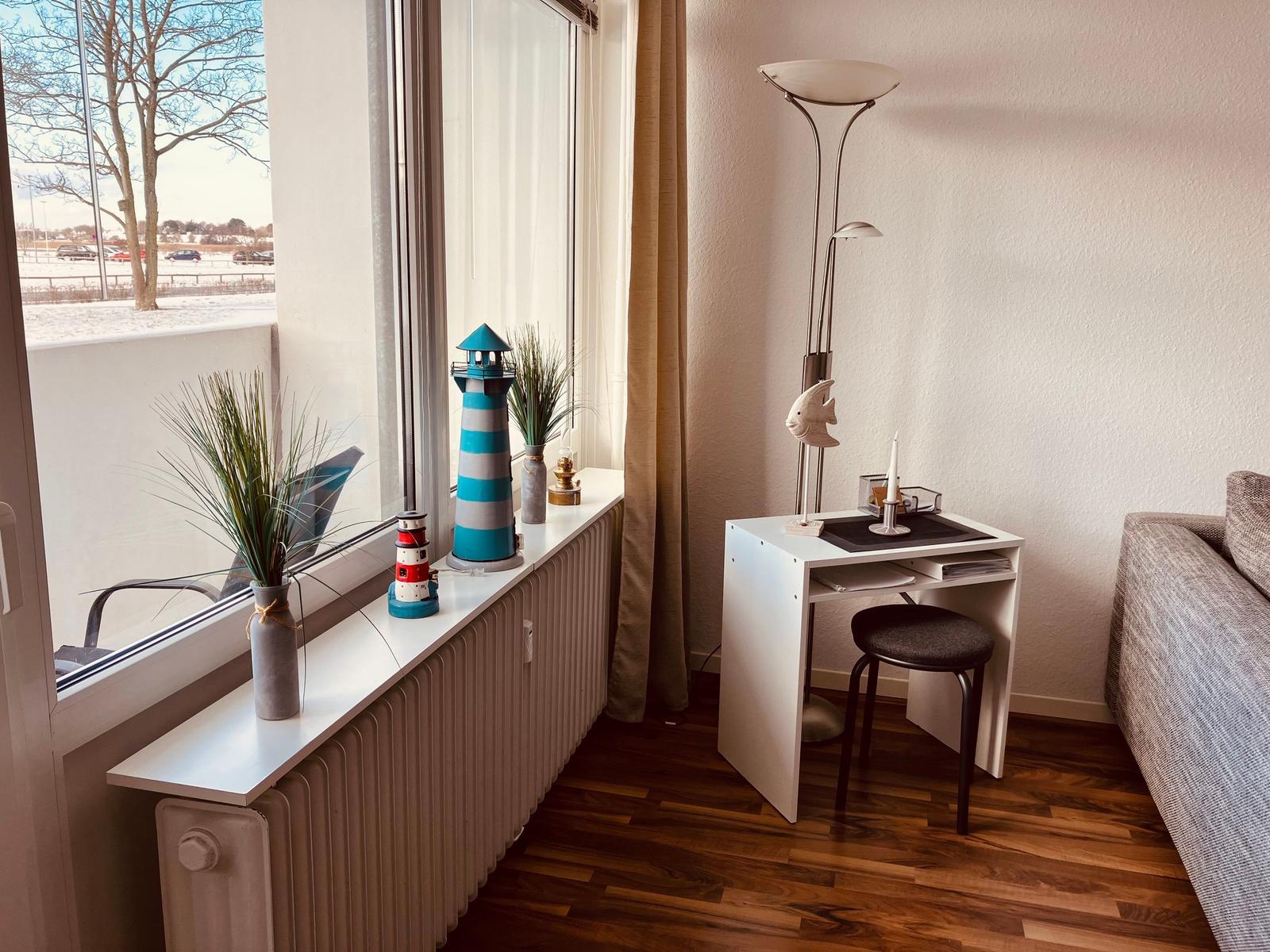 Ein kleiner Schreibtisch mit Stuhl steht neben einem Fenster. Auf dem Fensterbrett stehen Dekorationsgegenstände.