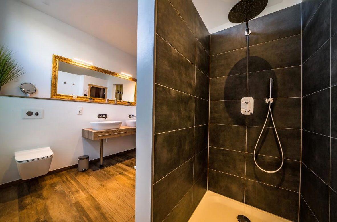 Dusche mit Duschbrause und Handbrause. Toilette und Waschbecken im angrenzenden Raum.