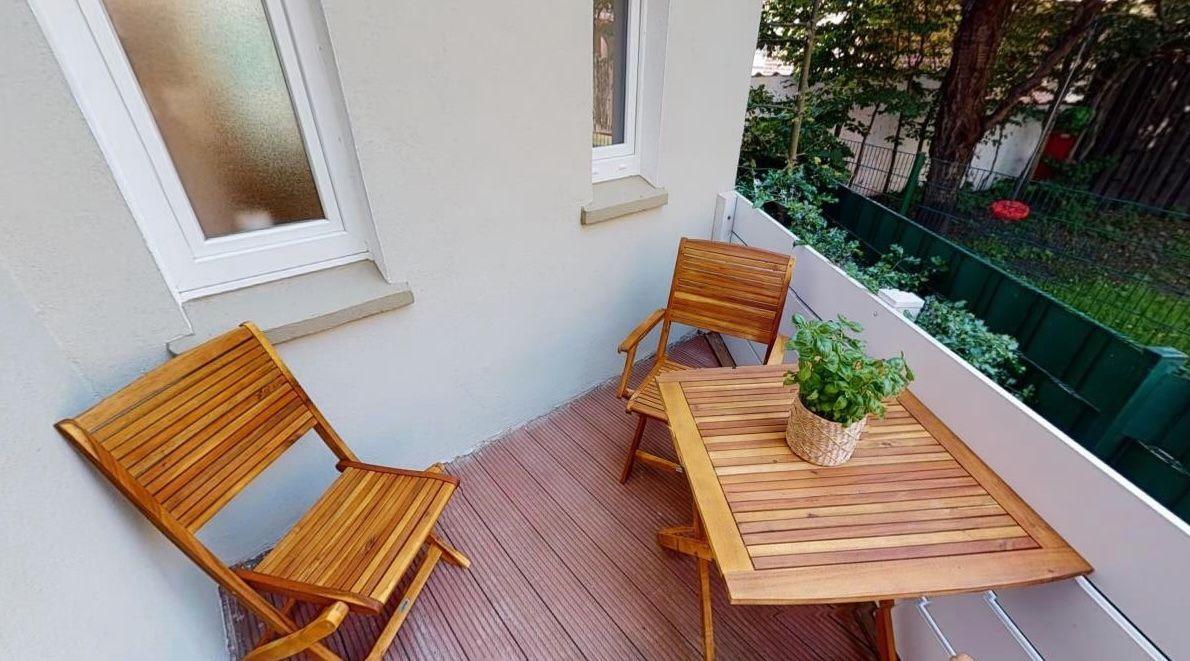 Terrasse mit Holztisch, zwei Stühlen und Pflanze. Hintergrund mit Grün und Zaun.