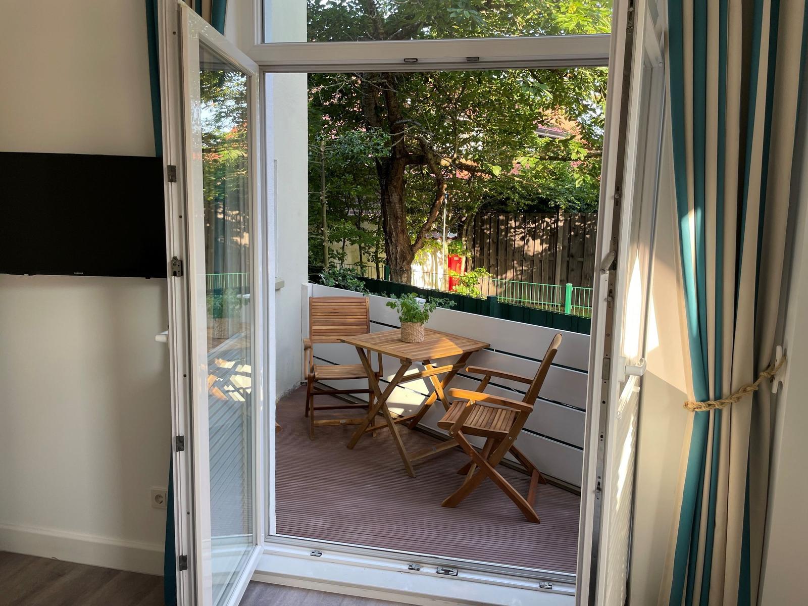 Terrasse mit Tisch und zwei Stühlen, umgeben von Bäumen und einem Zaun.