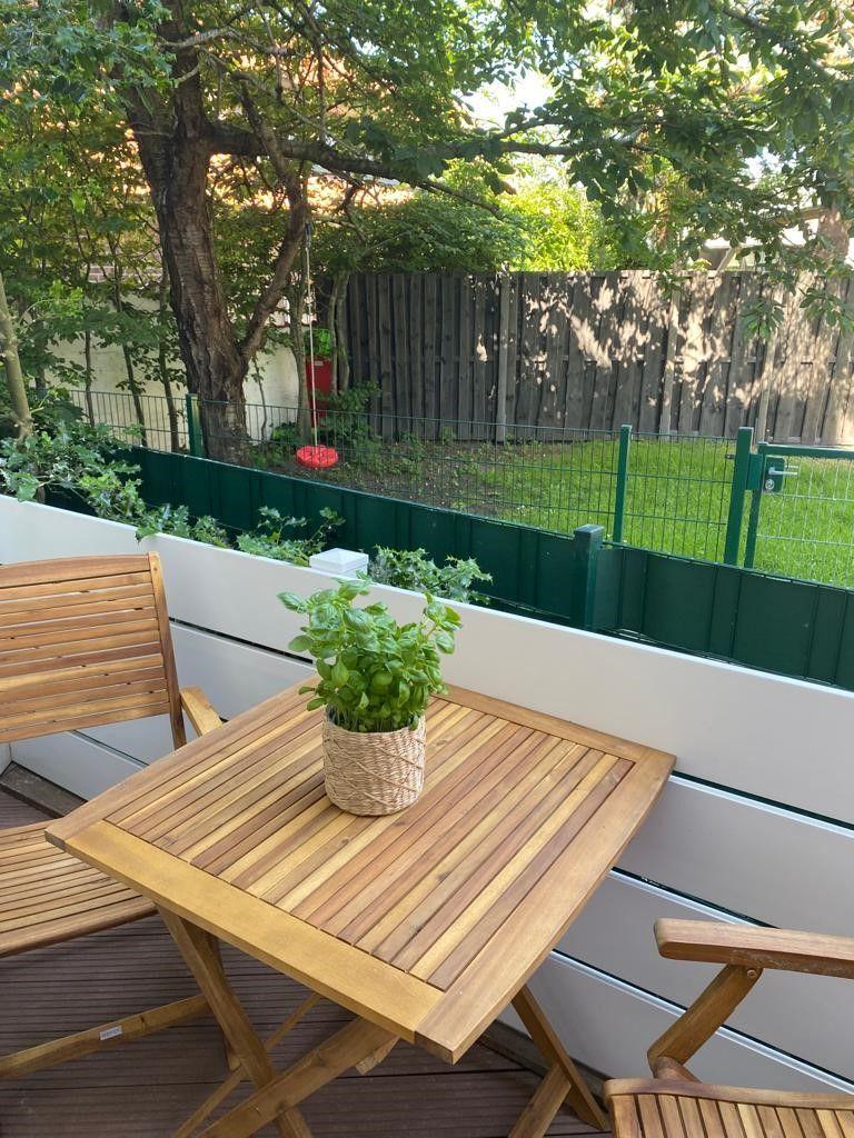 Terrasse mit Holztisch, zwei Stühlen und Pflanze. Hintergrund: grüner Zaun und Baum.