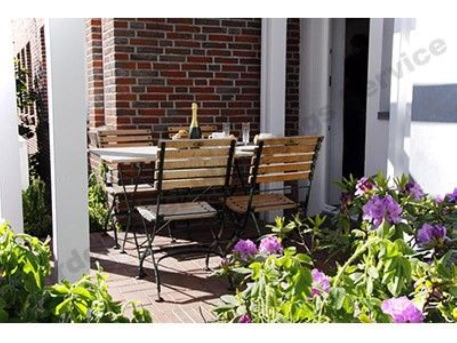 Terrasse mit Holzstühlen, Tisch und Flasche. Vorne Blumenbeet mit lila Blüten.