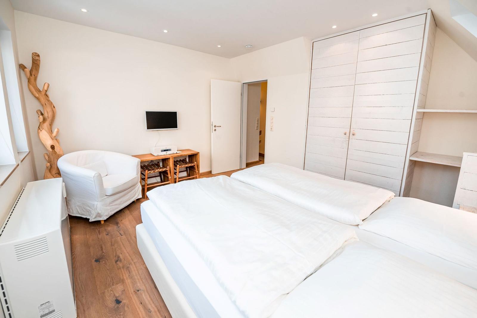 Zimmer mit Doppelbett, Kleiderschrank und Sessel. Holzboden und Wand-TV.