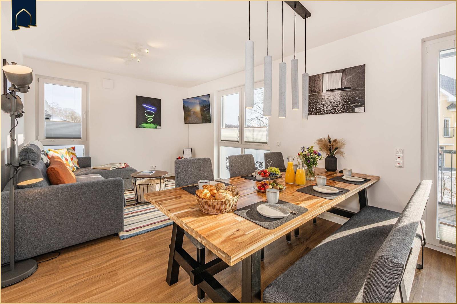 Essbereich mit Holztisch, Stühlen und Frühstück. Wohnbereich mit Sofa und Fenstern.