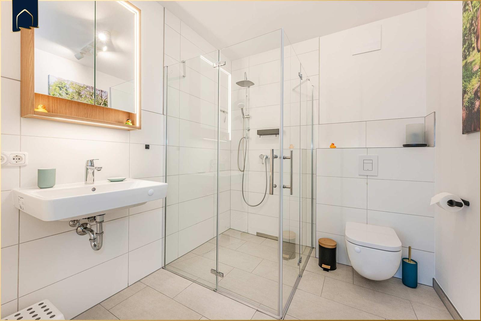 Badezimmer mit Dusche, Waschbecken und Toilette. Weiße Fliesen und Glasduschkabine.