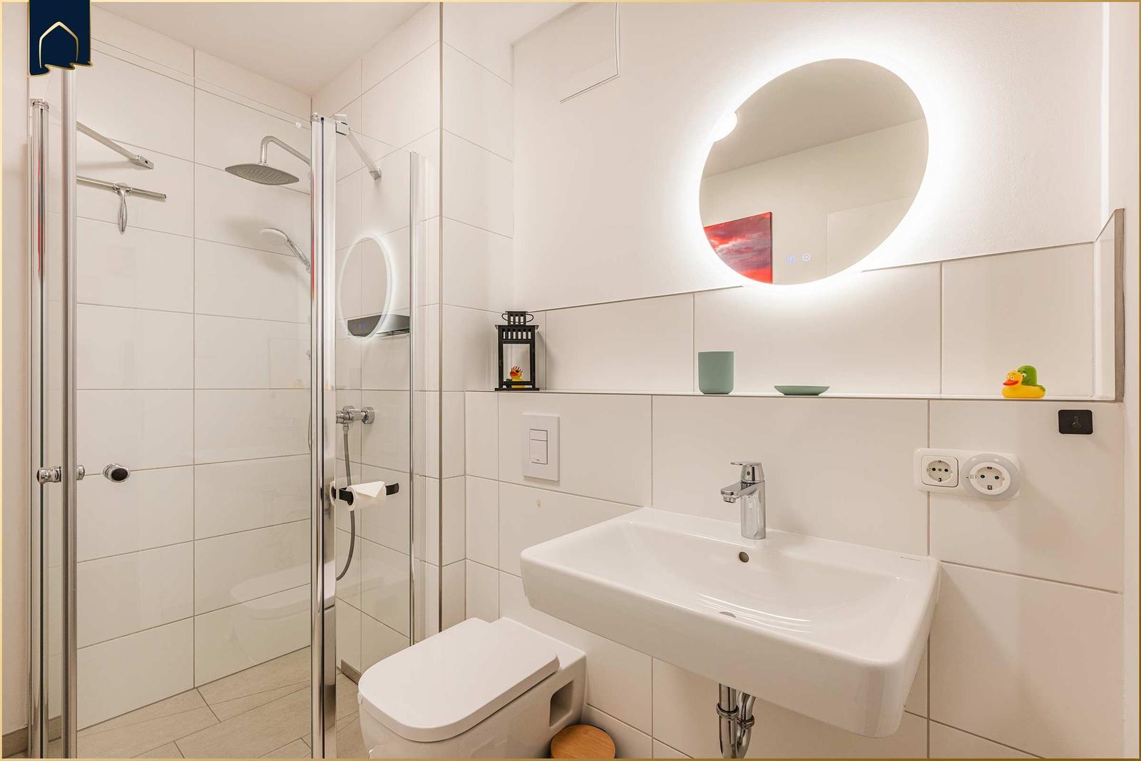 Badezimmer mit Dusche, Waschbecken und Toilette. Rundes Spiegel mit Beleuchtung.