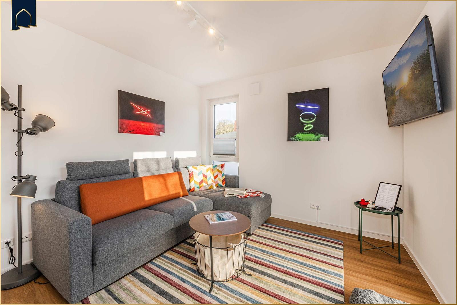 Ein Wohnzimmer mit grauem Sofa, bunten Kissen und Wandbildern.
