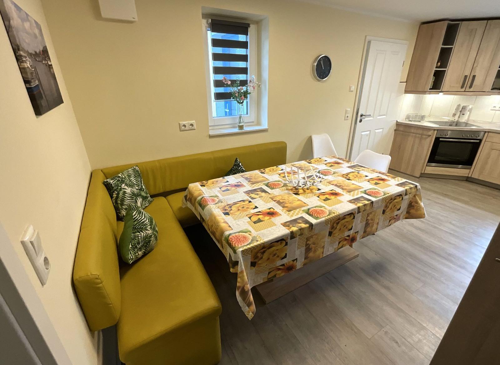 Gelber Sofa-Bereich neben Tisch mit Blumen-Tischdecke. Küche mit Holzschränken und Edelstahlherd.