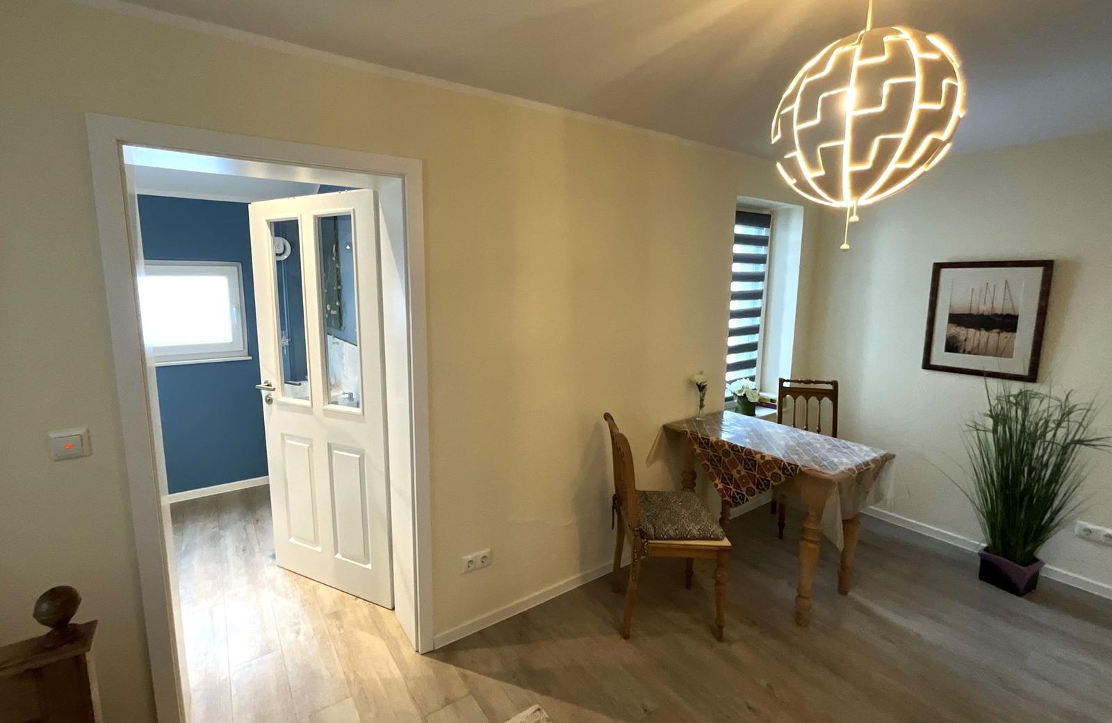 Esszimmer mit Tisch, Stühlen und Deckenleuchte. Tür führt in Schlafzimmer.