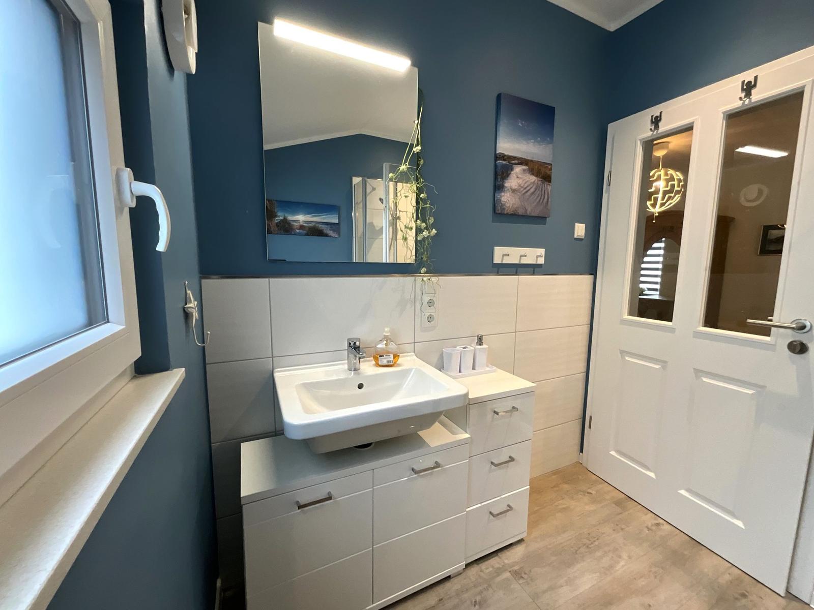 Badezimmer mit Waschbecken, Spiegel und Fenster. Wandfarbe blau, Boden hell.