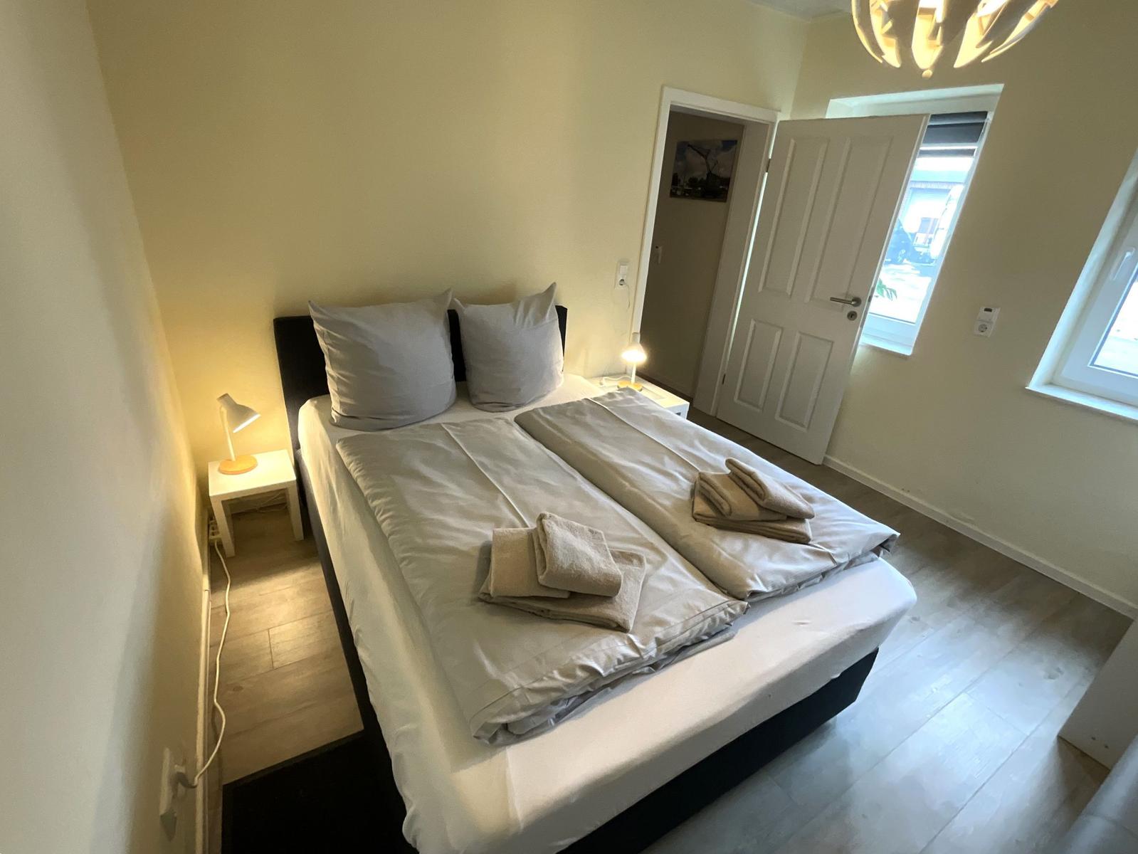 Schlafzimmer mit Bett, Nachttisch und Lampen. Tür führt zu einem Flur.
