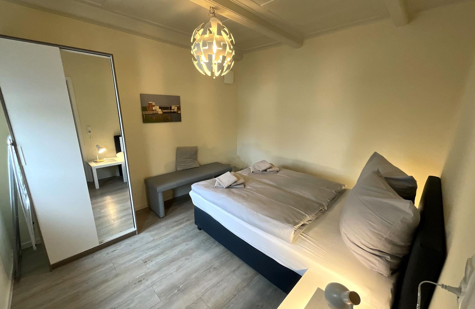 Schlafzimmer mit Bett, Kleiderschrank und Hängespiegel. Deckenleuchte und Holzboden.