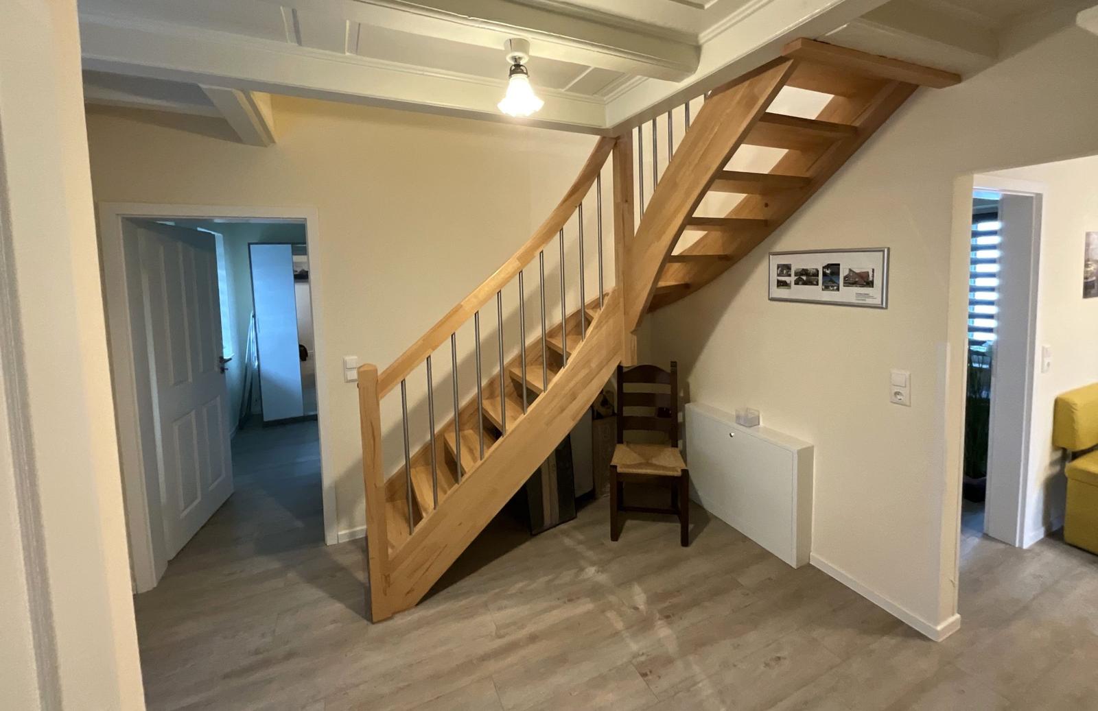 Holztreppenhaus mit Stufen und Geländer. Im Hintergrund sind Türen und ein Stuhl sichtbar.