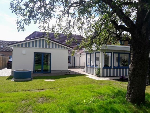 Haus mit Garten, blau-weißen Fenstern und großen Fenstern. Großer Baum im Vordergrund.