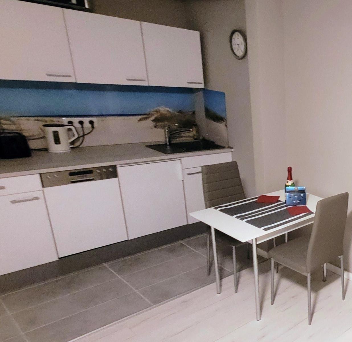 Küche mit Essbereich, weiße Schränke, Tisch mit Stühlen und Wanduhr.