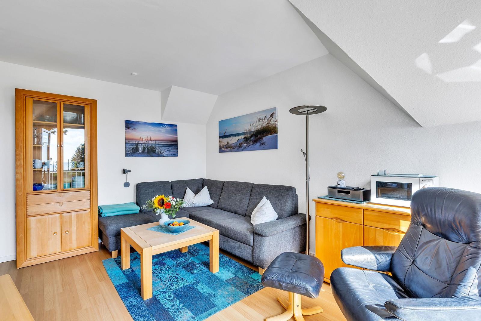 Wohnzimmer mit Sofa, Sessel und Küchenschränkchen. Blauer Teppich und Wandbilder.