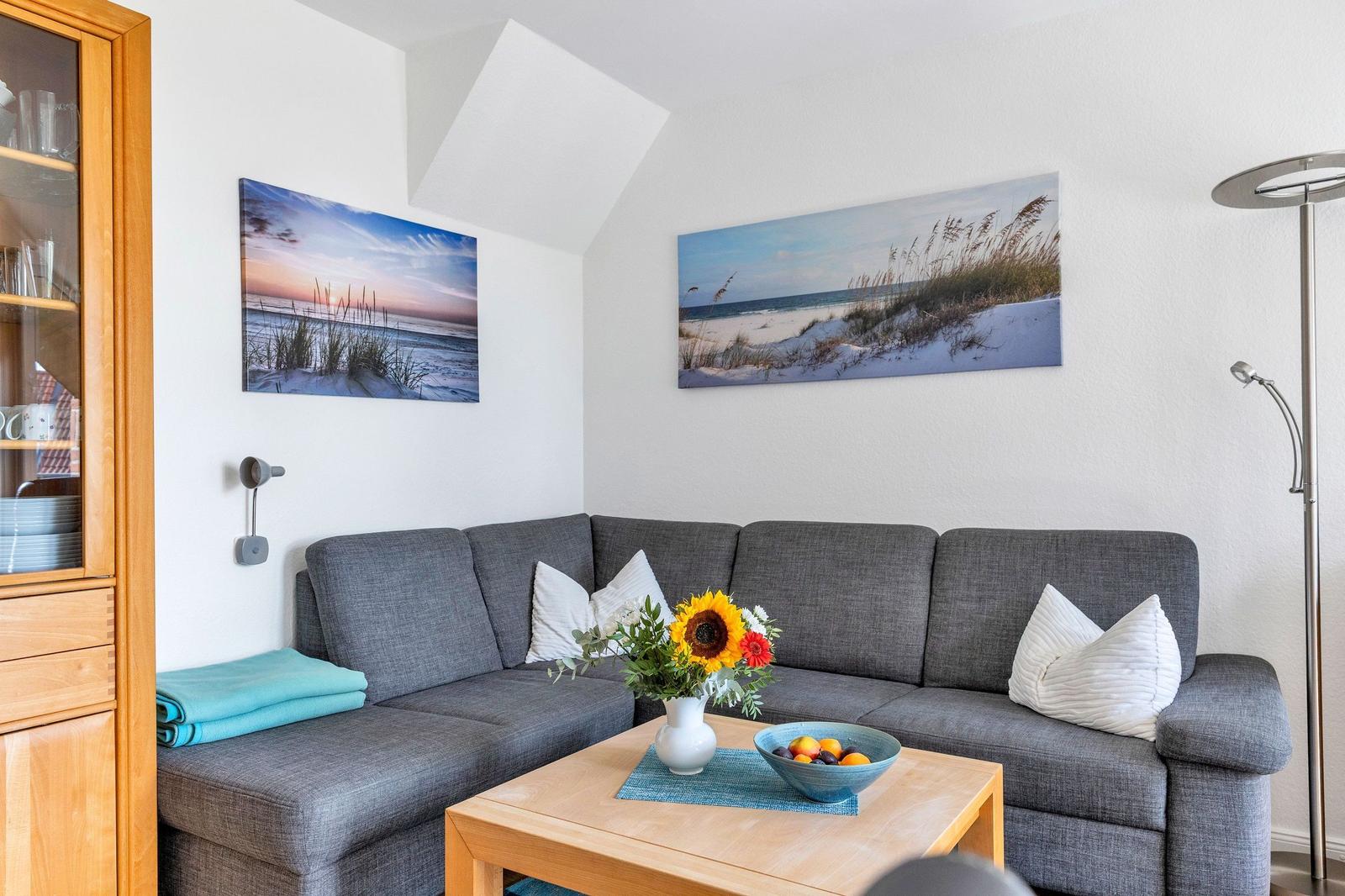 Grauer Sofa mit weißen Kissen, Holztisch, Wandbilder und Stehlampe.