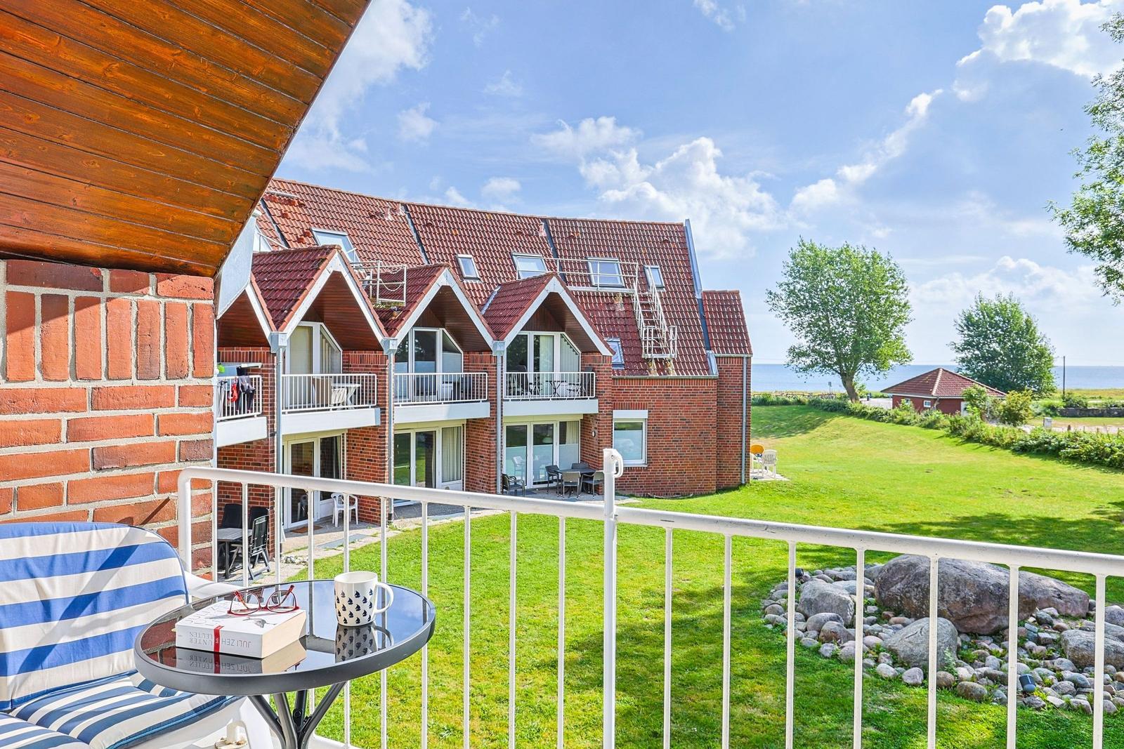 Terrasse mit Tisch und Stuhl. Blick auf roten Backsteinbau mit Balkonen und grünen Wiesen.