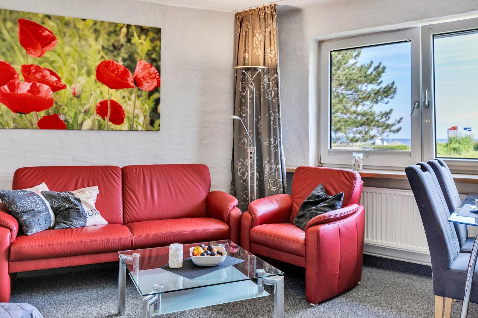 Wohnzimmer mit rotem Sofa, Sessel und Glas-Tisch. Fenster mit Blick auf Bäume.