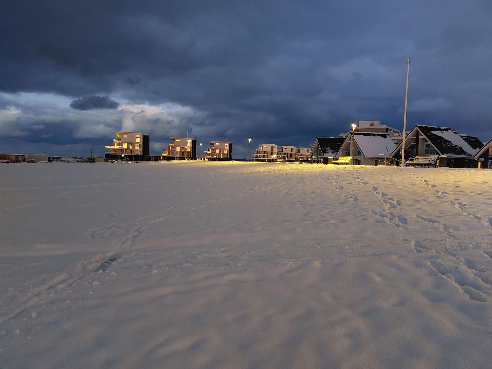 Schneebedeckter Platz mit beleuchteten Häusern unter bewölktem Himmel.