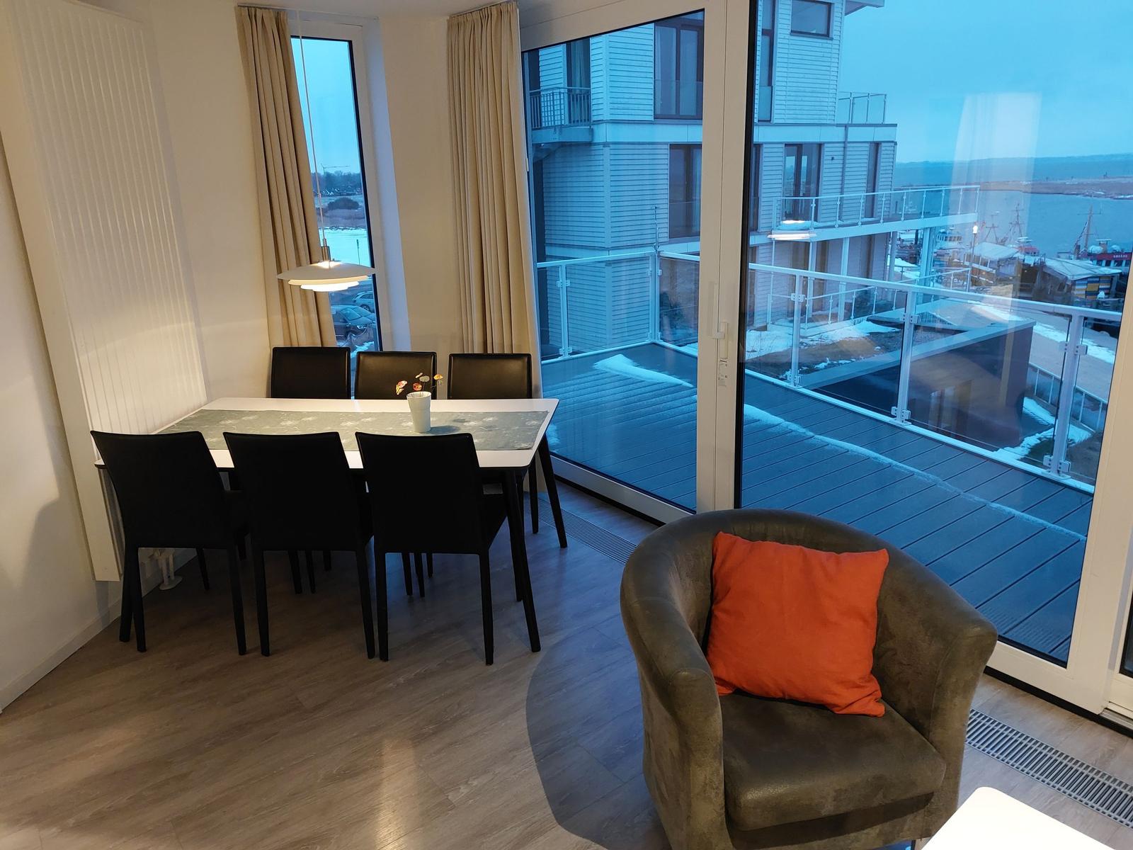 Essbereich mit Tisch und Stühlen, Sessel mit Kissen, Blick auf Terrasse und Hafen.