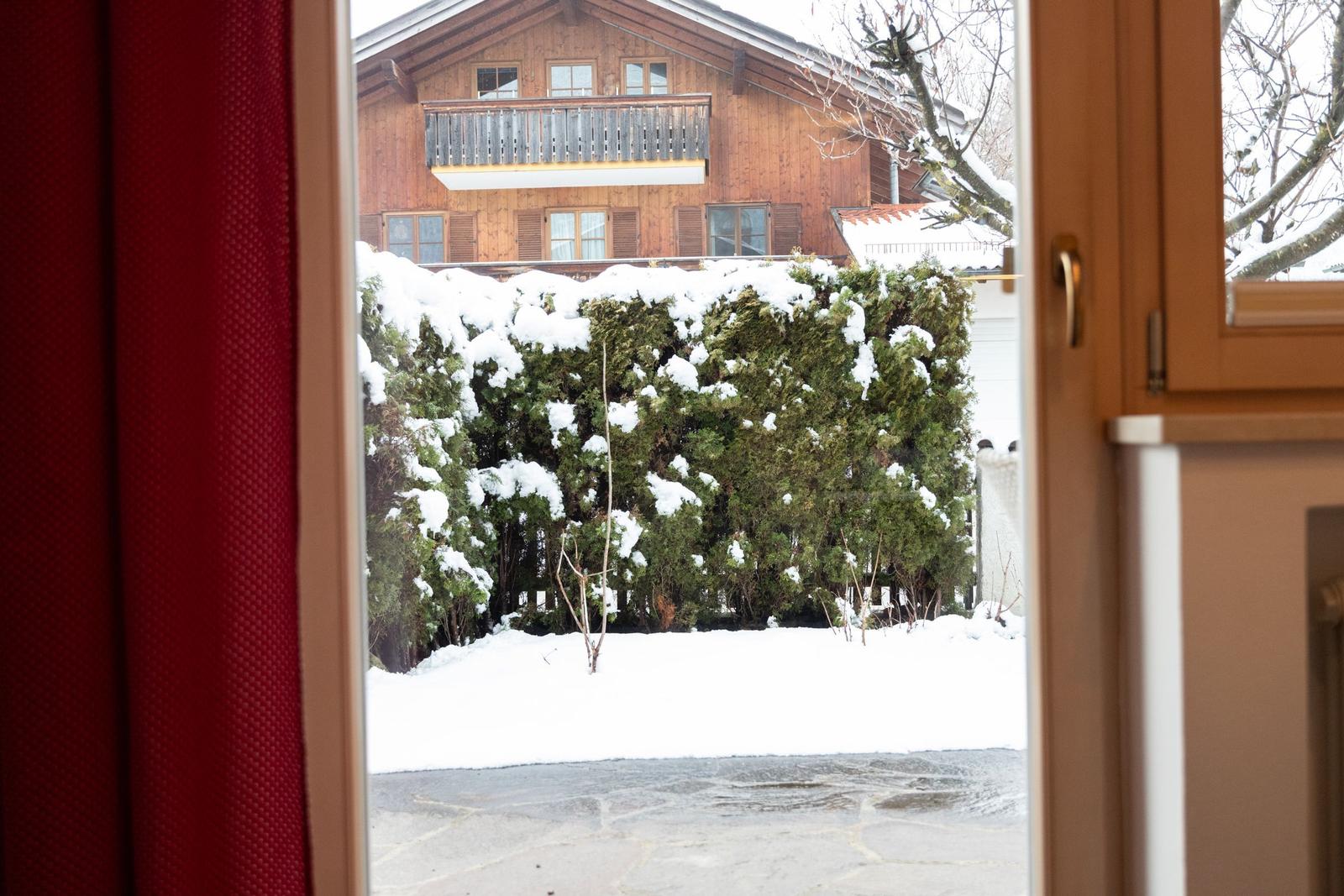 Durch das Fenster blickt man auf einen schneebedeckten Garten mit Hecken und ein Holzhaus.