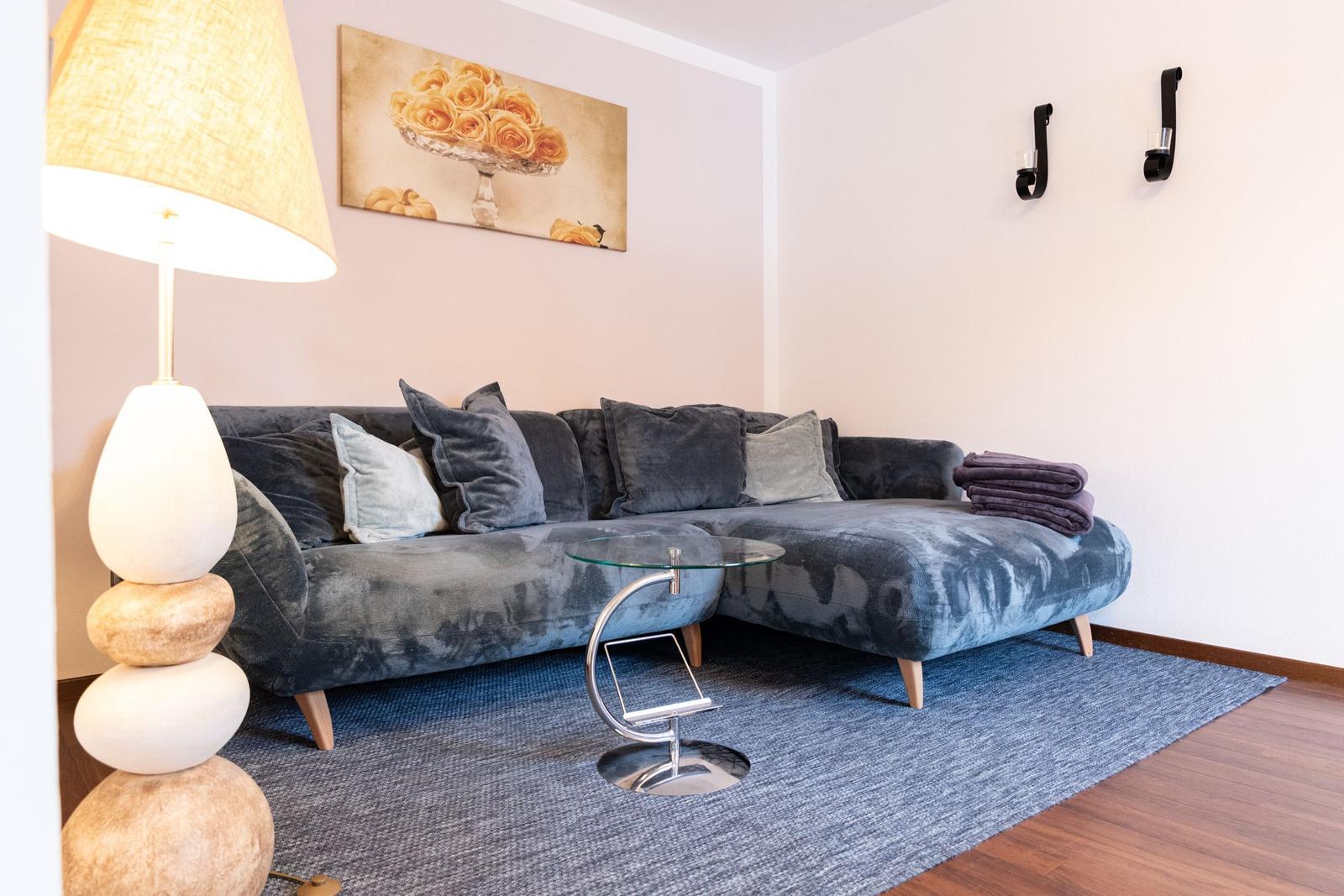 Ein Wohnzimmer mit einem grauen Sofa, einem Glas-Tisch und einer Stehlampe.