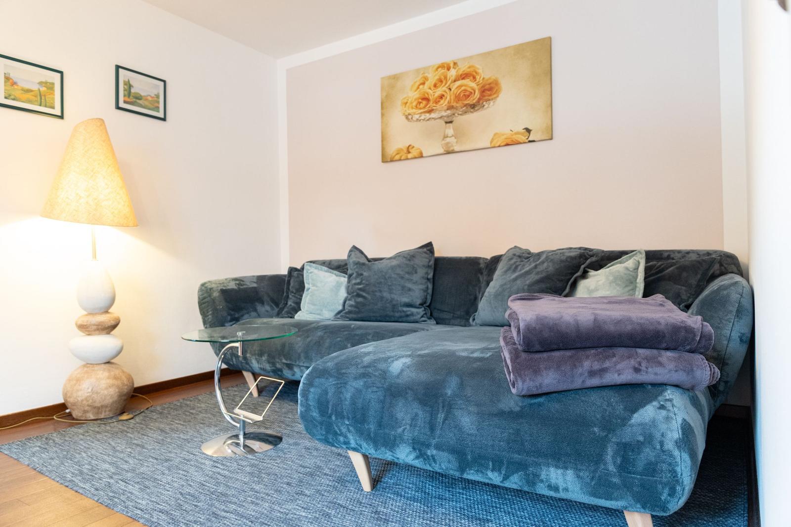 Ein Wohnzimmer mit blauem Sofa, Glas-Tisch und Wandbild mit Rosen.