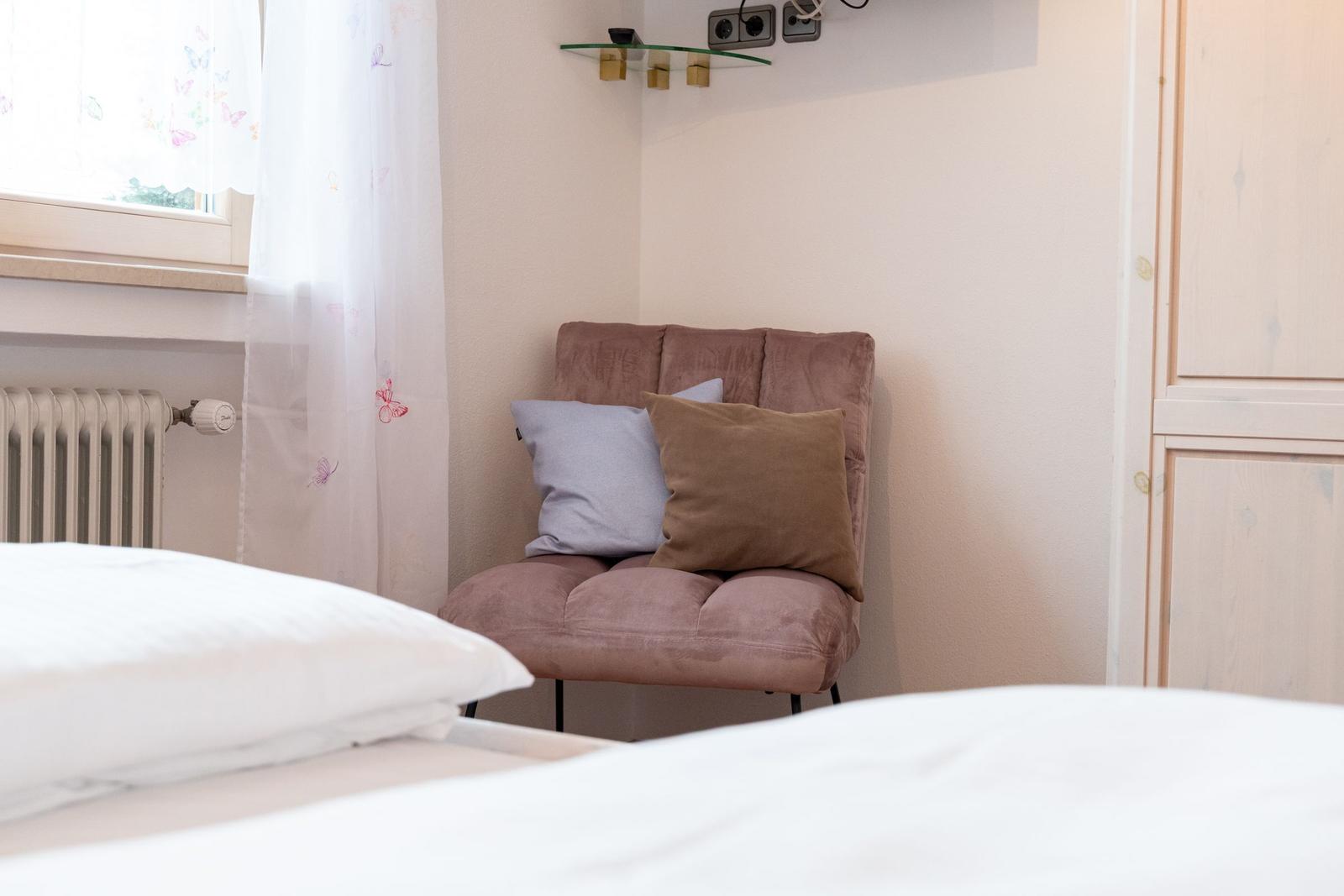 Ein Bett mit weißer Bettwäsche, ein brauner Sessel mit Kissen und ein Fenster mit weißen Vorhängen.