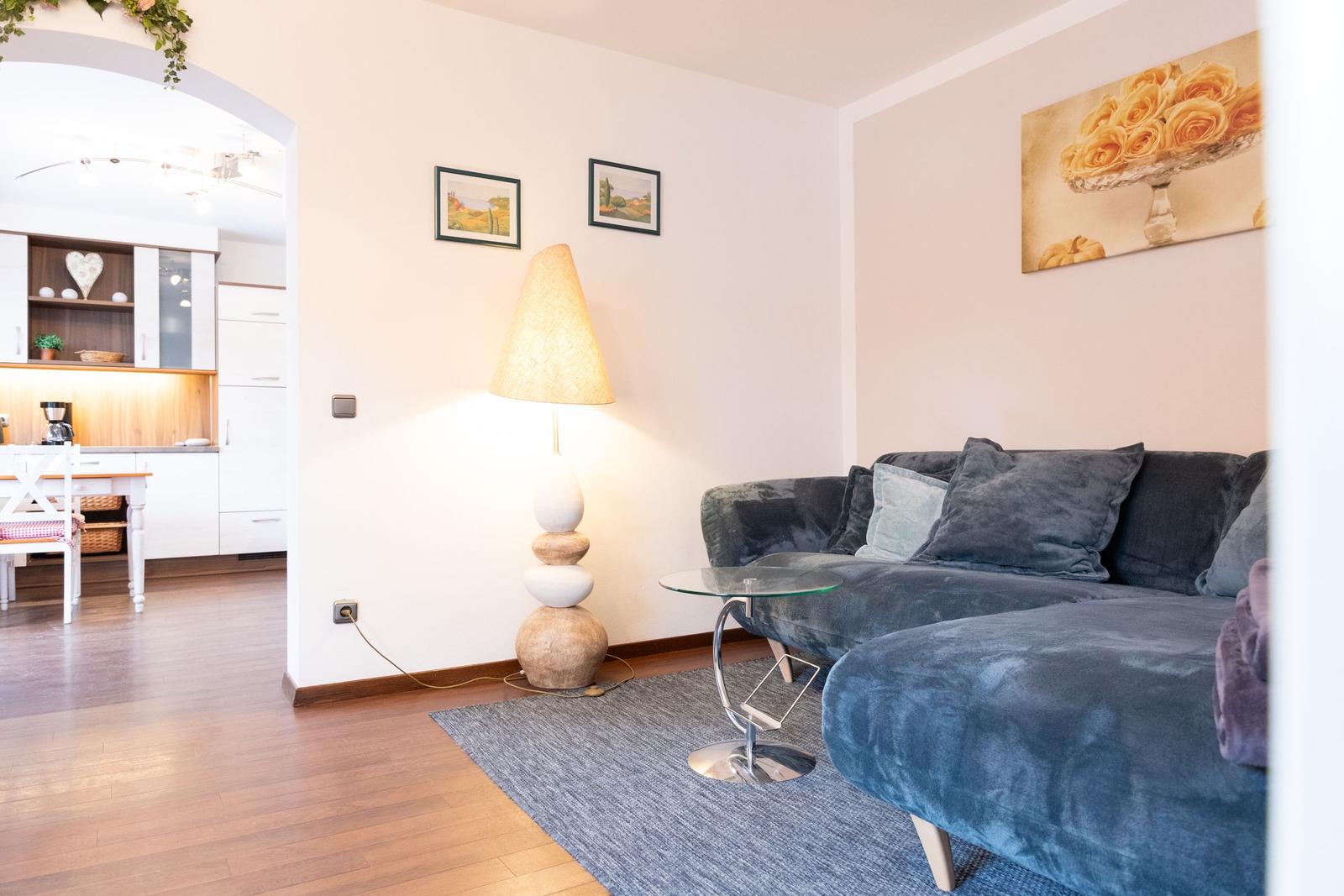 Wohnzimmer mit Sofa, Stehlampe und Glas-Tisch. Küche im Hintergrund sichtbar.