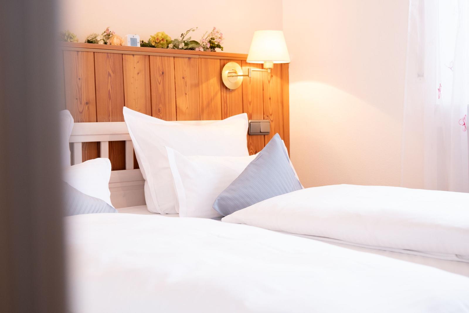 Doppelbett mit weißer Bettwäsche und Holzkopfteil. Wandlampe und Blumen dekorieren den Raum.
