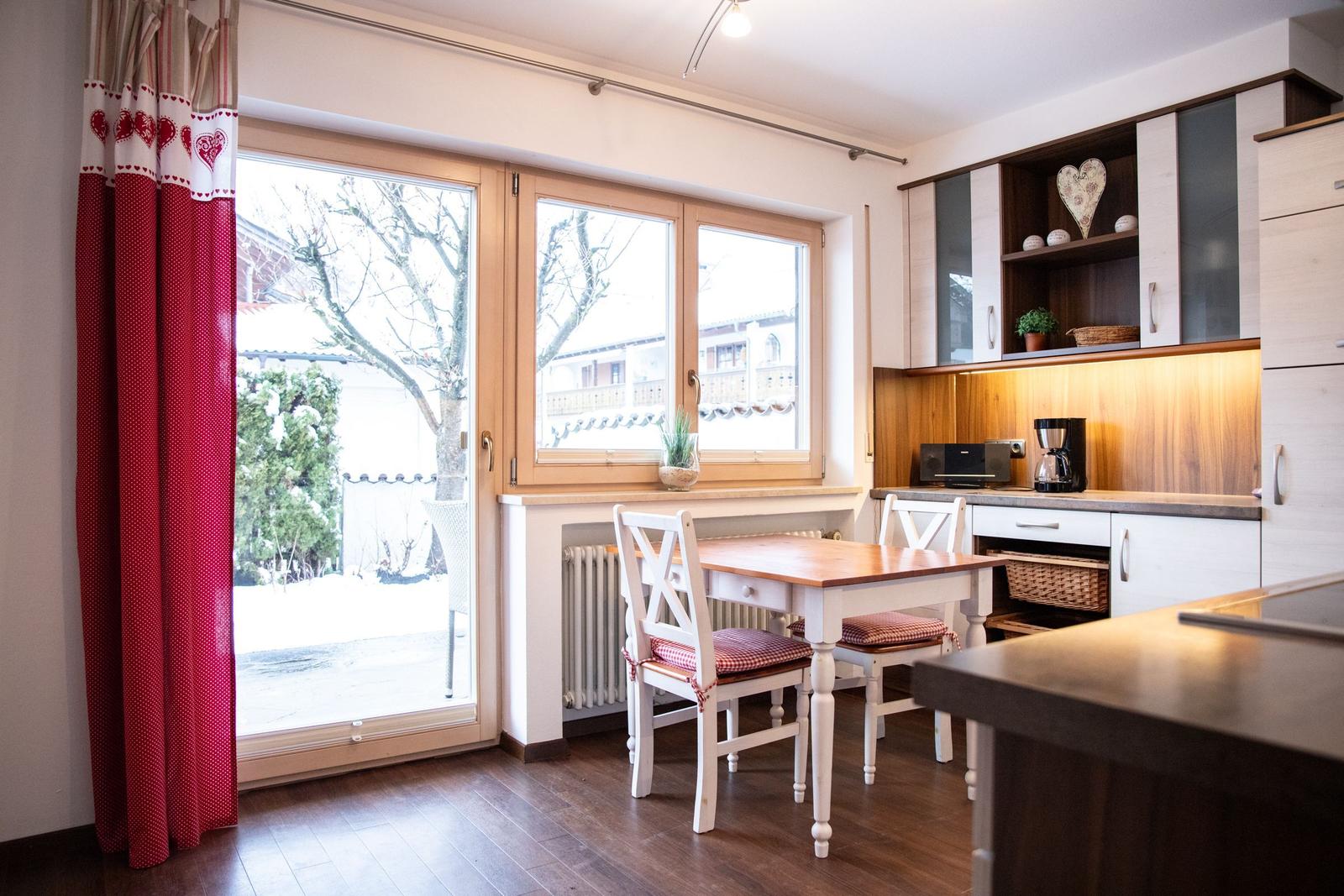Essbereich mit Tisch, Stühlen und Küchenzeile. Fenster mit roten Vorhängen. Draußen Schnee.
